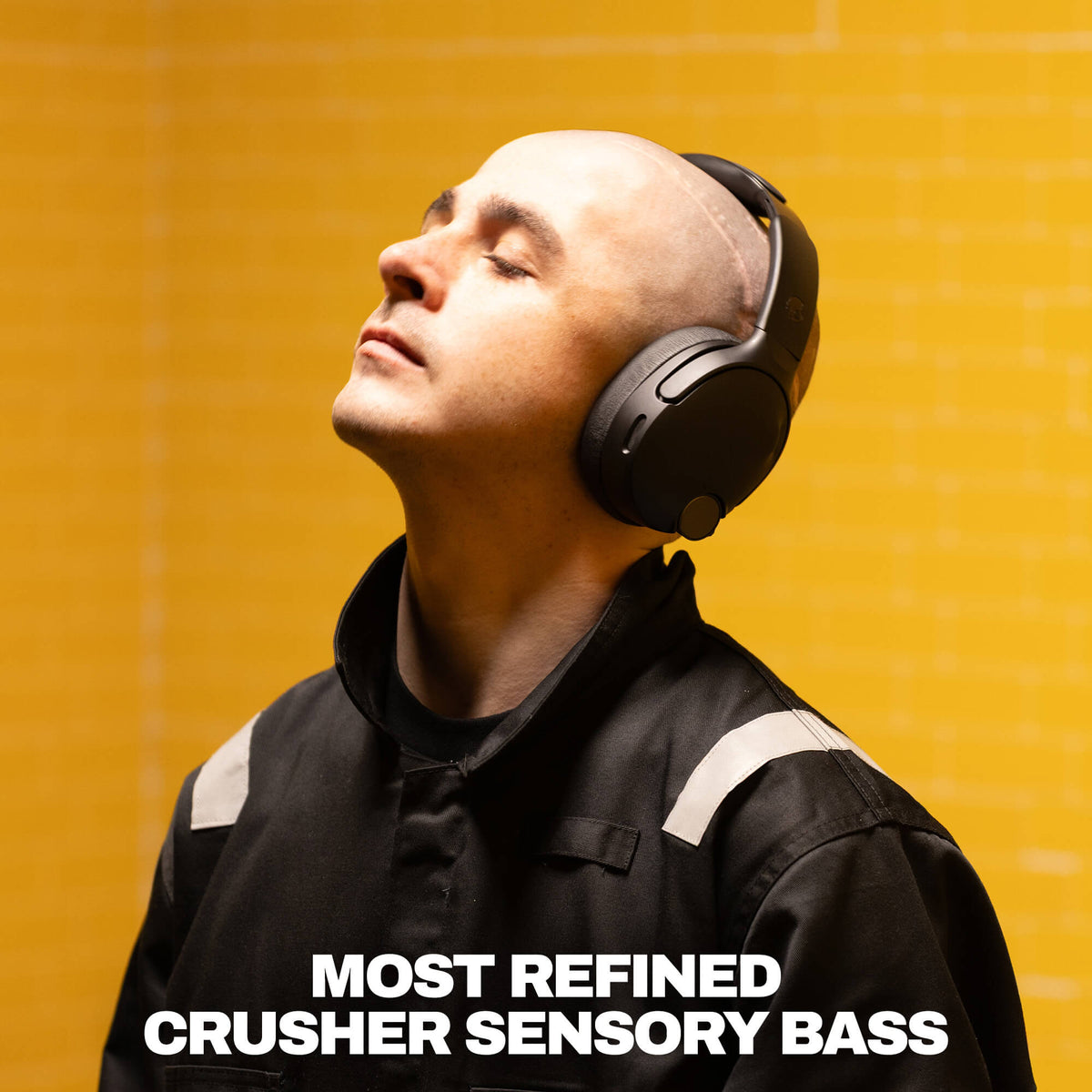 True Black | A man wearing black over-ear headphones and a black jacket with white shoulder stripes, looking upward against a bright yellow background. Large white text at the bottom reads 'MOST REFINED CRUSHER SENSORY BASS'.