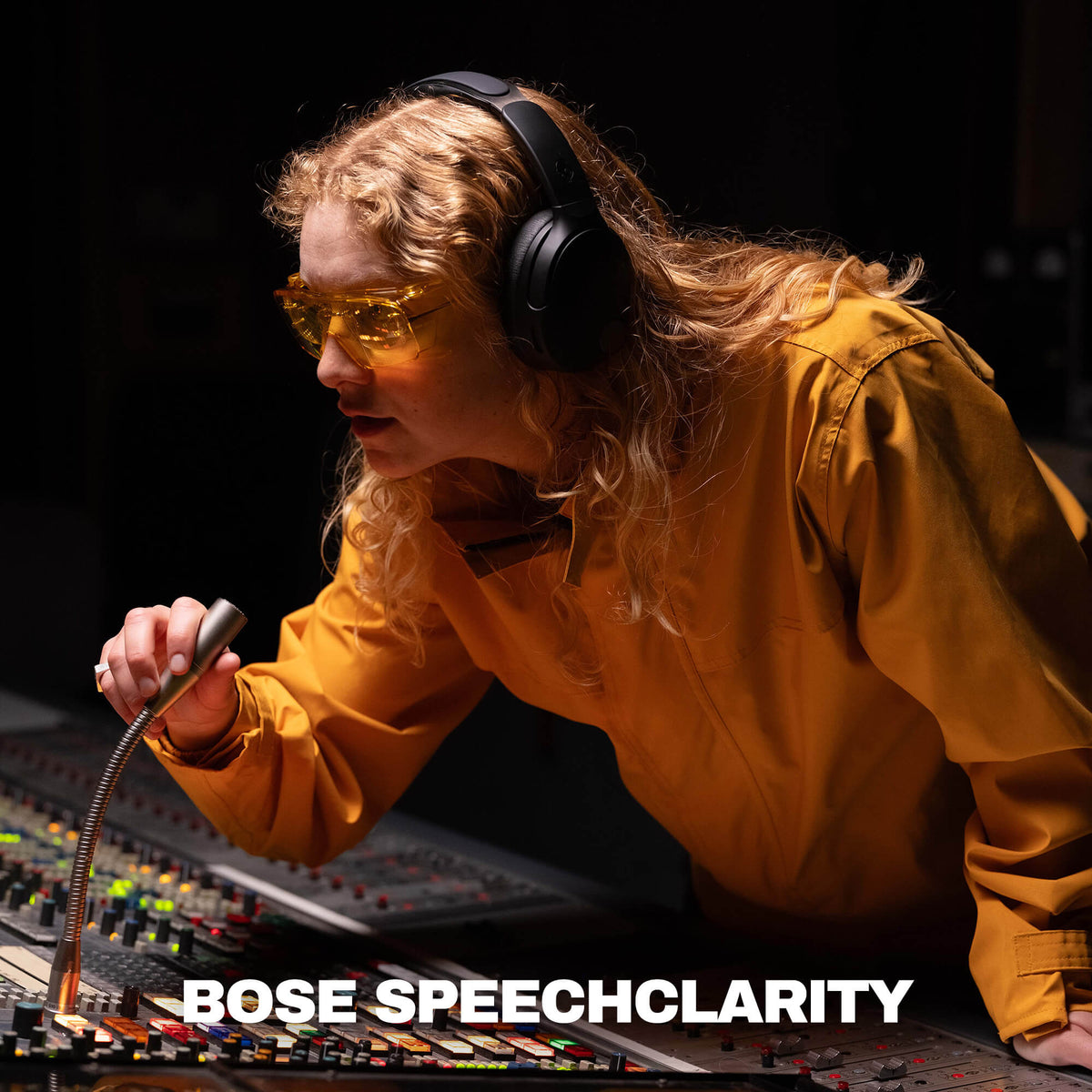 True Black | A woman in a recording studio wearing yellow-tinted safety glasses and black Crusher 1080 over-ear headphones, leaning over a mixing board and holding a microphone. The text 'BOSE SPEECHCLARITY' appears at the bottom.