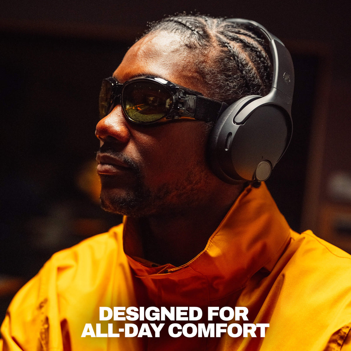 True Black | A man in a bright orange jacket wearing black over-ear headphones and dark sunglasses, shown in a dim indoor setting with the text 'DESIGNED FOR ALL-DAY COMFORT' at the  bottom.