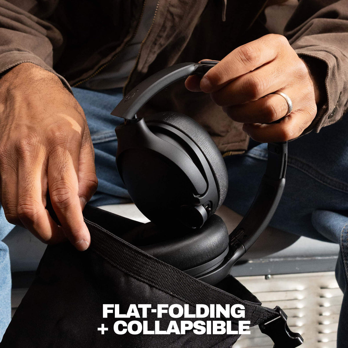 True Black | A close-up of a person holding a black over-ear headset over an open black bag, with the text 'FLAT-FOLDING + COLLAPSIBLE' at the bottom. The person is wearing a brown jacket and blue jeans.