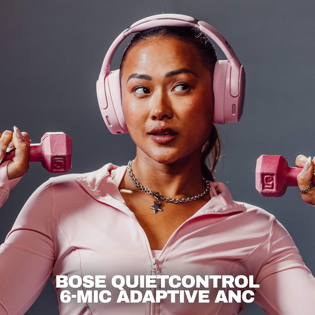 Candy | A woman wearing pink Crusher 1080 headphones with Bose QuietControl 6-MIC Adaptive ANC over-ear headphones and a matching pink athletic zip-up top, holding pink dumbbells on both sides against a dark gray studio background. White text at the bottom reads 'BOSE QUIETCONTROL 6-MIC ADAPTIVE ANC'.