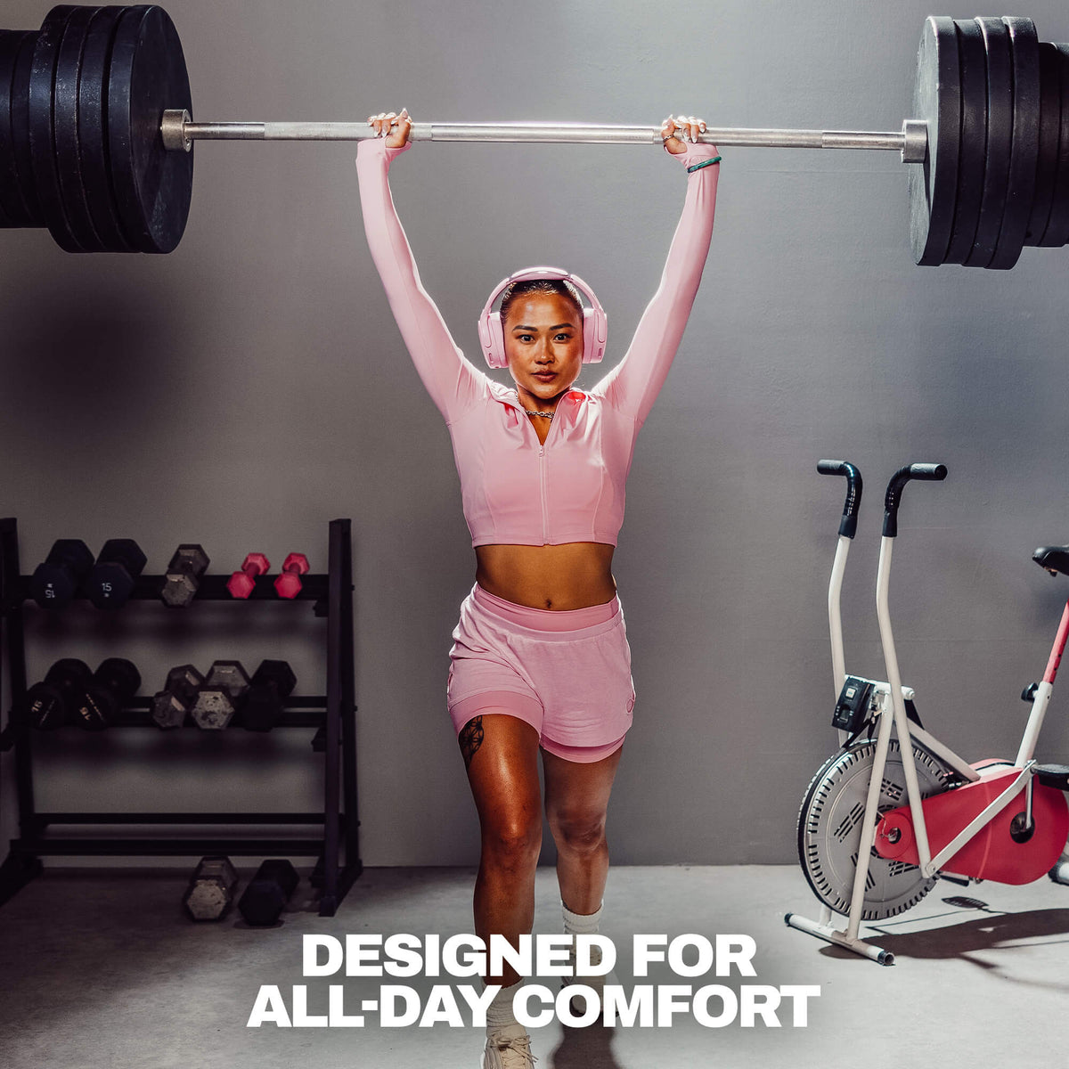 Candy | A woman in a pink athletic zip-up hoodie and matching shorts lifts a barbell overhead in a gym setting. She is wearing pink headphones, with dumbbells on a rack to her left and a stationary bike to her right.