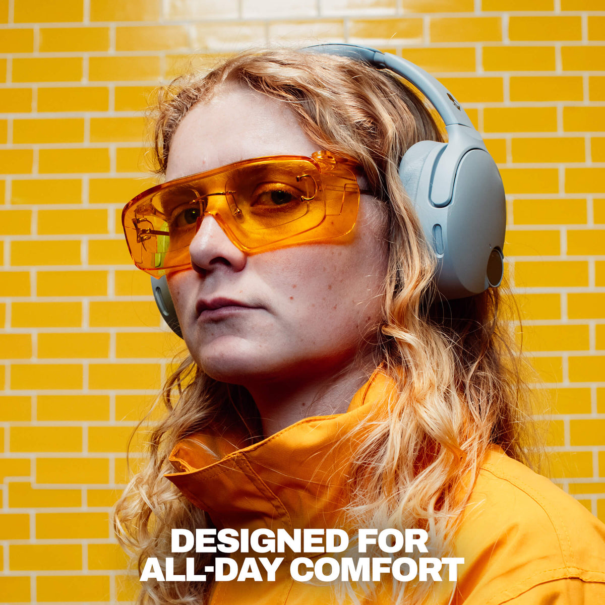 Cement | A woman wearing crusher 1080  over-ear headphones and translucent orange-tinted safety glasses, set against a yellow brick wall background with the text 'DESIGNED FOR ALL-DAY COMFORT' at the bottom.