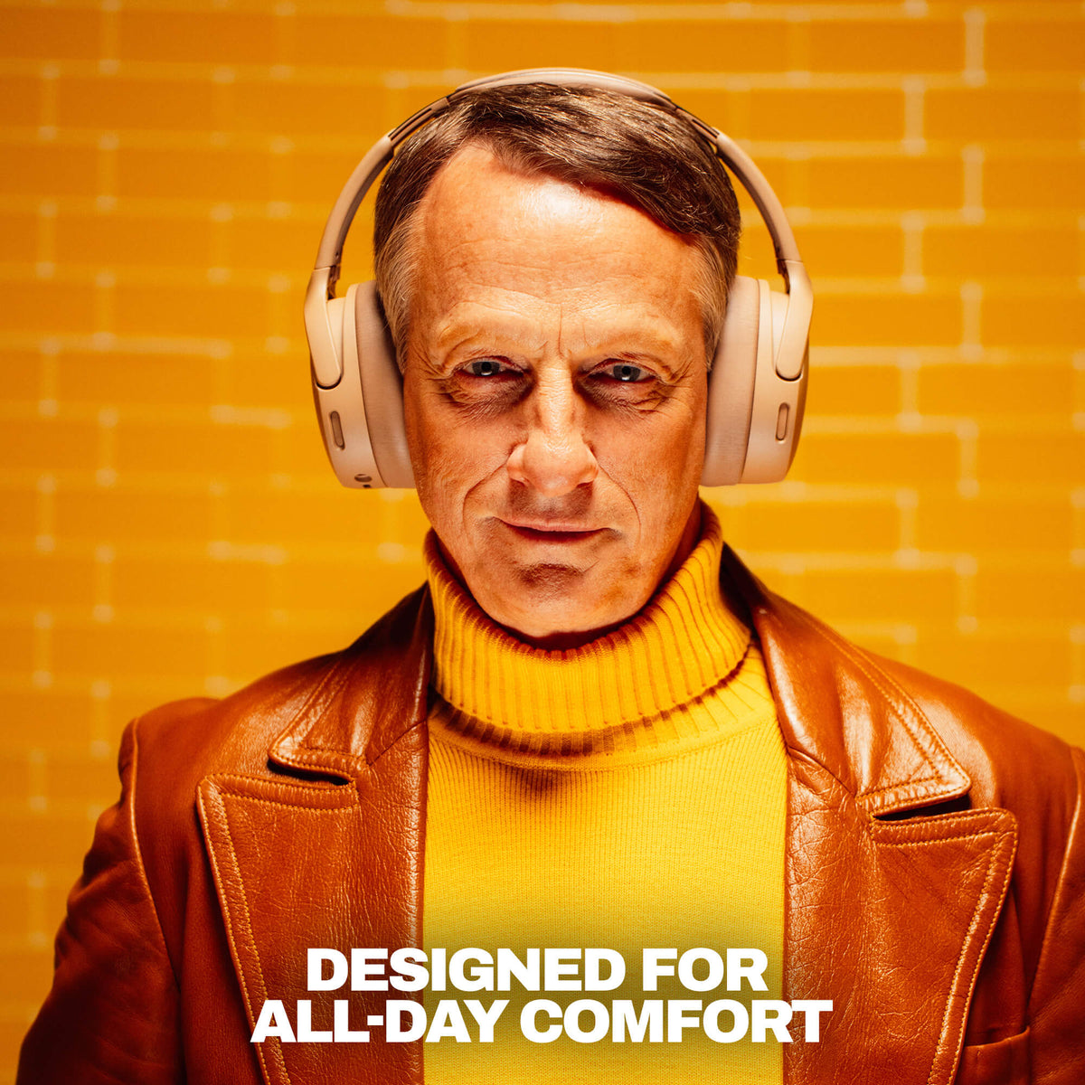 Primer | tony hawk wearing beige crusher 1080 over-ear headphones, a brown leather jacket, and a yellow turtleneck sweater stands against a yellow brick wall background with the text 'DESIGNED FOR ALL-DAY COMFORT' at the bottom.