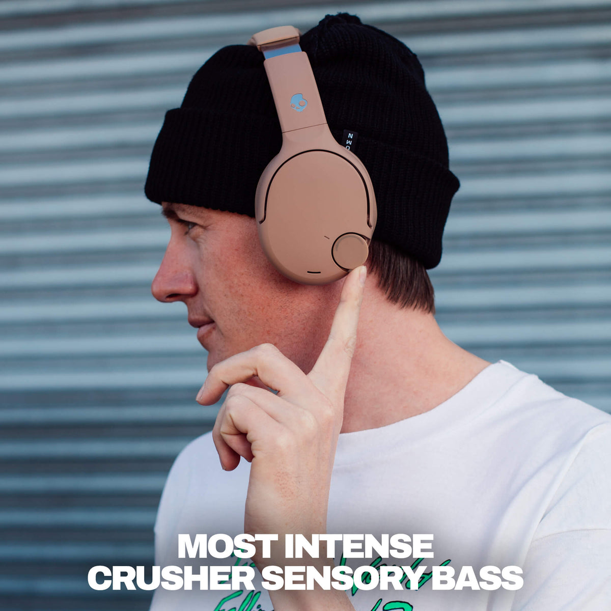 Primer | Man wearing Crusher 720 headphones over a black beanie looking sideways and 'MOST INTENSE CRUSHER SENSORY BASS' text against a stripe textured blue metal wall.