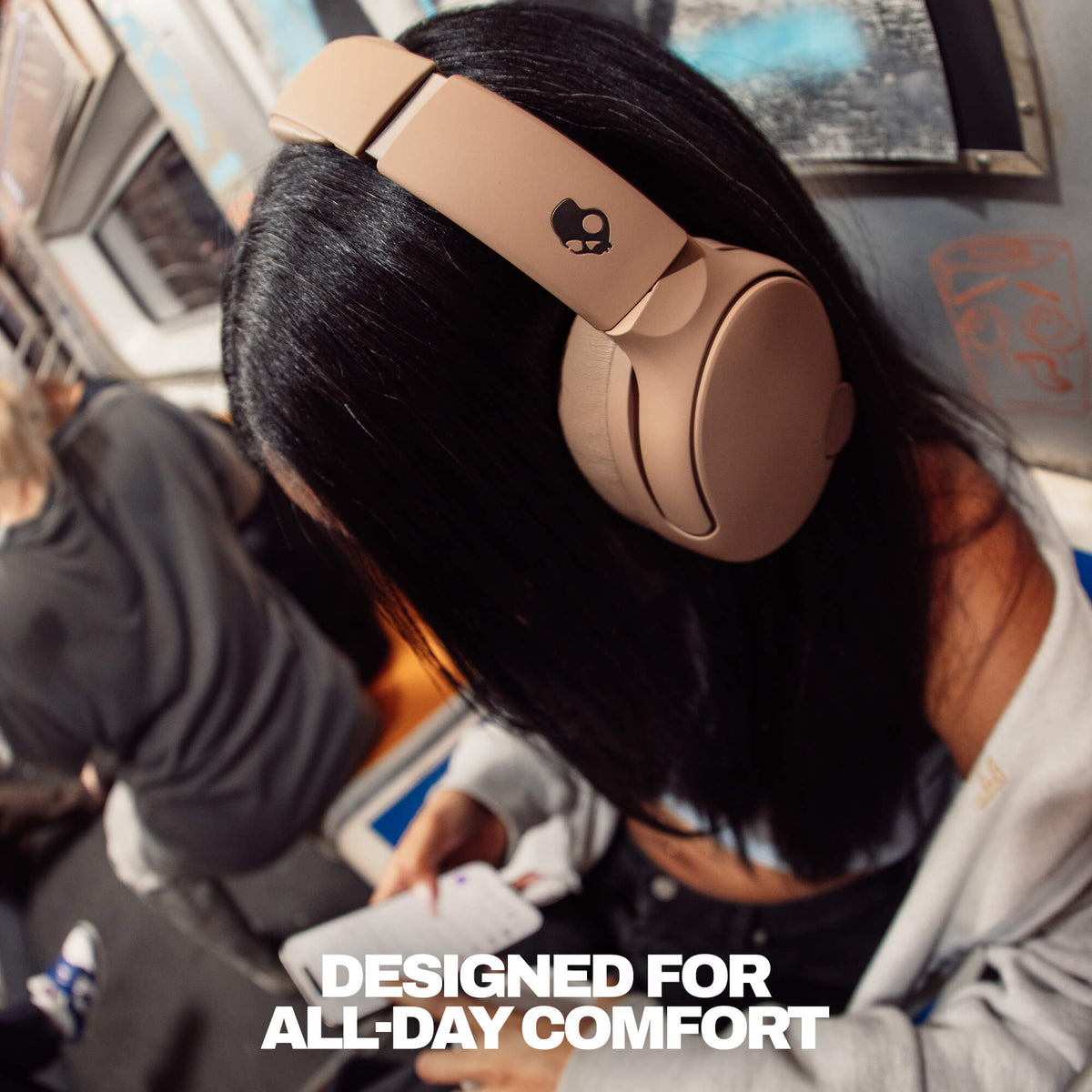 Primer | A person with long black hair wearing beige over-ear headphones on a subway train, with text 'DESIGNED FOR ALL-DAY COMFORT'.